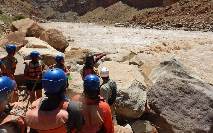 A group stands on the shore of a river while an instructor points out over the water